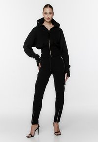 Czarna kurtka z kapturem krótką, z szerokimi rękawami, połączona z czarnymi spodniami cargo z zamkami na kostkach i dyskretnymi kieszeniami bocznymi.