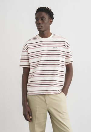 Hombre con pantalones beige y una camisa de manga corta blanca con finas rayas horizontales marrones y rosas y un pequeño logo de "Dickies".