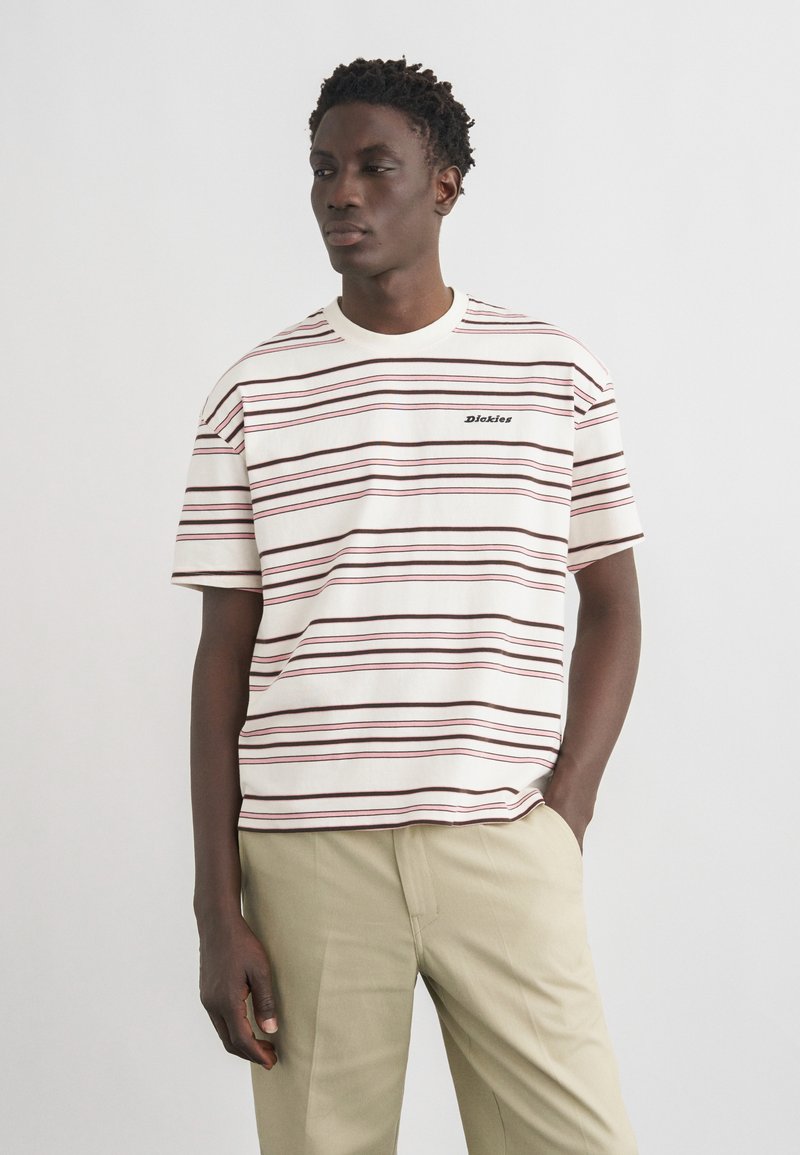 Hombre con pantalones beige y una camisa de manga corta blanca con finas rayas horizontales marrones y rosas y un pequeño logo de "Dickies".