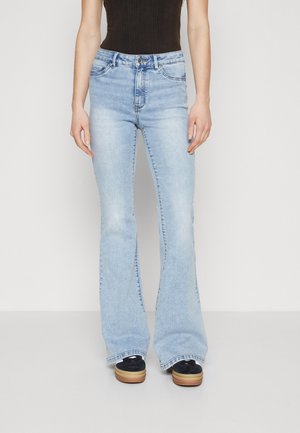 Personne portant un jean évasé bleu clair et des chaussures à plateforme noires, debout devant un fond clair uni.