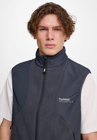 Junger Mann mit lockigem braunem Haar, der eine marineblaue ärmellose Zip-Jacke über einem weißen Kurzarmhemd trägt und nach vorne schaut.