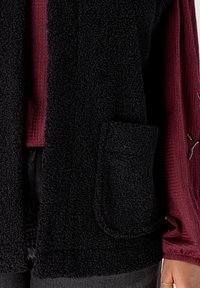 Zwarte, fuzzy sjaalvest met een zak, gedragen over een bordeaux ribbeltop, die de textuur van de stof en het kleurcontrast benadrukt.