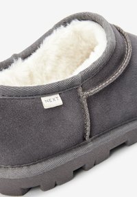 Pantofole in camoscio grigio con fodera in pelliccia sintetica, collo arrotondato e suola in gomma texturizzata; presenta un'etichetta visibile con il nome del marchio "NEXT".