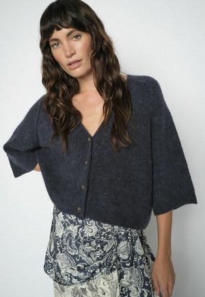 Frau mit langen, welligen braunen Haaren trägt eine dunkelgraue, flauschige, geknöpfte Strickjacke und einen gemusterten Rock mit Paisley-Motiven.