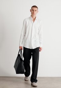 Chemise blanche à boutons avec manches longues, associée à un pantalon noir et à un sac fourre-tout noir. Les chaussures sont des baskets grises. Fond neutre.