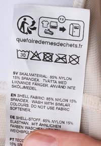 Étiquette d'entretien avec composition du tissu : 85 % nylon, 15 % élasthanne. Instructions d'entretien en plusieurs langues. Texte : fond blanc, police noire.