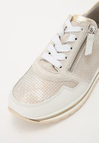 Sneaker con una toma in metallo intrecciato beige, dettagli in pelle bianca, lacci bianchi e una cerniera laterale. Superfici texture e lisce.