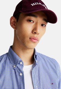 Camisa de cuadros azul y blanca con botones y cuello, superpuesta sobre una camiseta blanca, combinada con una gorra burdeos con letras blancas.