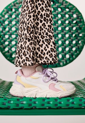 Personne portant un pantalon imprimé léopard et des chaussettes roses côtelées, debout sur une chaise tissée verte, portant des baskets épaisses aux couleurs pastel.