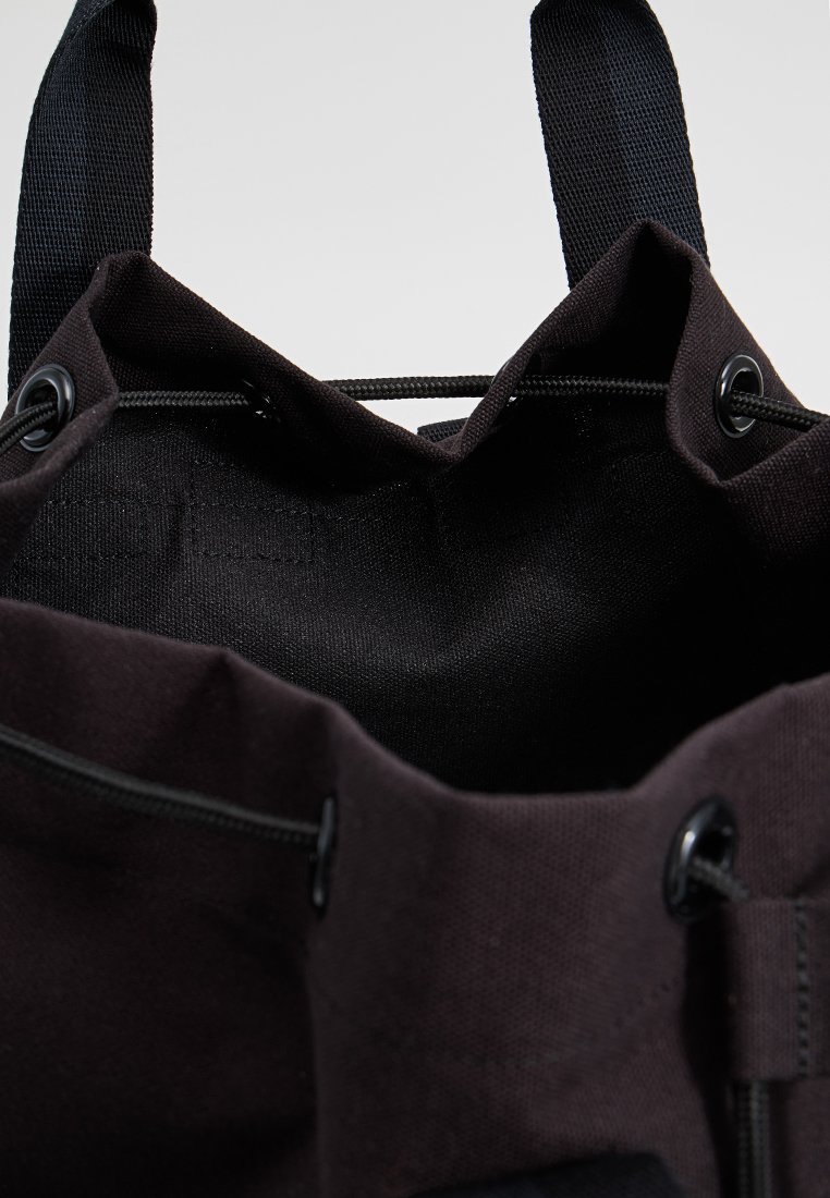 Bolsa de lona negra con una abertura redonda, que cuenta con un cierre ajustable de cordón y mangos de cinta resistentes. El interior tiene una textura lisa y mate.