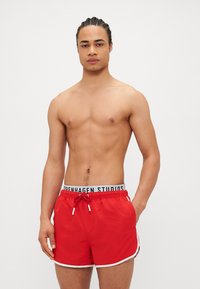 Red swim shorts with a white trim, elastic waistband featuring "Copenhagen Studios" logo, and adjustable drawstring. Smooth fabric texture.