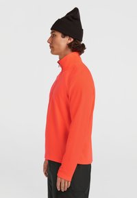 Pomarańczowy polarowy sweter z wysokim kołnierzem i zapinanym przodem, w połączeniu z czapką beanie z czarnej dzianiny; gładka tekstura, dopasowany krój.