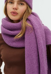 Ung kvinde iført en brun top, lilla strikhue og matchende tyk lilla strikscarf viklet omkring halsen.