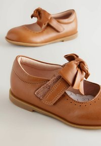Braune Lederschuhe mit runder Zehenpartie, Riemenschluss und Schleifendetail. Mit belüfteter Zehenpartie und strukturierten Akzenten am Riemen.