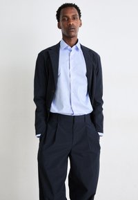 Navy blazer over light blue striped shirt with a collar, paired with dark tailored trousers featuring a loose fit and front pleats.