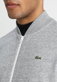 Světle šedá mikina na zip s žebrovaným límcem, na hrudi je malé zelené logo krokodýla a texturovaná tkanina.