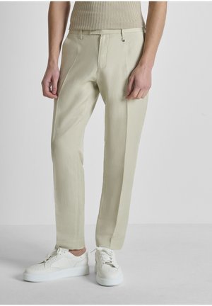 Pantalones a medida beige combinados con zapatillas blancas con cordones y una camiseta sin mangas de canalé beige, modelo posando contra un fondo liso.