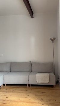 Γκρι γωνιακός καναπές με τρία μαξιλάρια και μπεζ κουβέρτα σε μινιμαλιστικό δωμάτιο με ξύλινο δάπεδο και ψηλό μεταλλικό φωτιστικό δαπέδου.