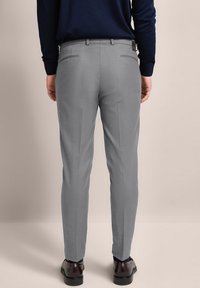 Graue Hose mit maßgeschneidertem Schnitt, ausgestattet mit Seitentaschen und Gesäßtaschen, kombiniert mit dunklen Schuhen. Glattes Material und Falten-Details.