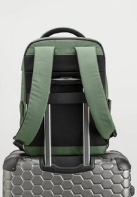 Mochila verde y negra con correas acolchadas y paneles de malla, colocada sobre una maleta plateada con patrón hexagonal.
