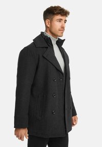 Schwarzer doppelt-knöpfbarer Peacoat mit breitem Kragen, Seitentaschen und einer glatten Textur. Getragen über einem grauen Rollkragenpullover.