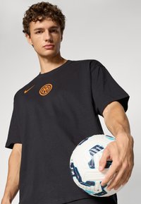 Musta puuvillapaita, jossa on oranssi logo rinnassa, päällä henkilö, joka pitää kädessään teksturoitua valkoista ja sinistä jalkapalloa.