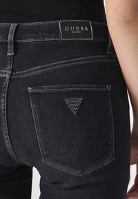 Jeans en denim noir avec un patch logo Guess, un triangle décoratif sur la poche arrière et des coutures contrastantes le long des coutures.