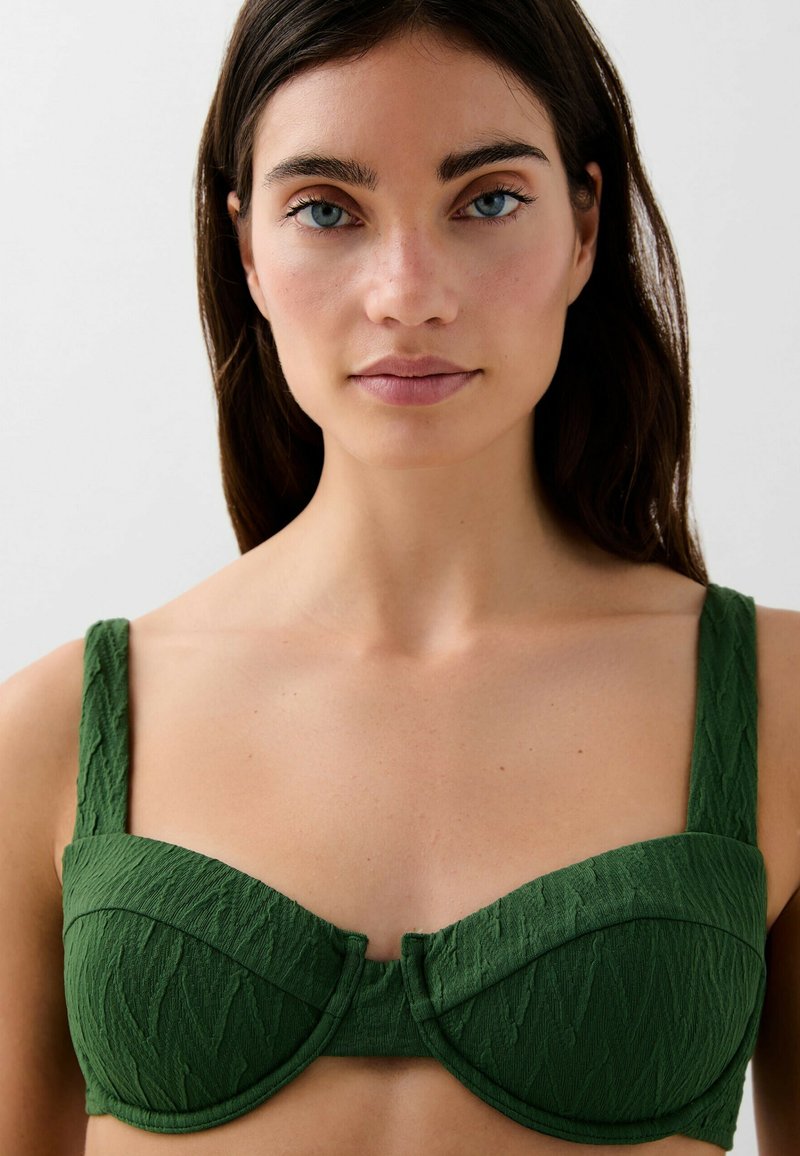 Joven mujer con ojos azules y cabello oscuro, vistiendo un top de sujetador sin tirantes de color verde oscuro y texturizado, mirando de frente contra un fondo liso.