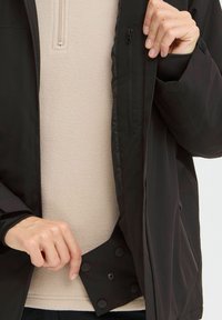 Svart jakke med et mykt beige fleecefôr. Har en glidelåslomme og trykknapper nederst. Glatt tekstur med kontrasterende materialer.