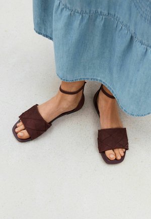 Feet wearing dark brown quilted sandals with ankle straps, shown with a light blue denim skirt on a light floor.