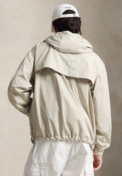 Homme portant une veste à capuche beige et un pantalon cargo blanc, de dos, avec une casquette de baseball Polo blanche sur fond neutre.