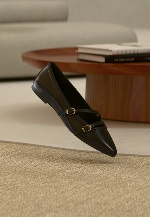 Chaussure plate noire à bout pointu avec deux fines brides à boucle flottant au-dessus d'un tapis beige, avec une table basse en bois et des livres en arrière-plan.