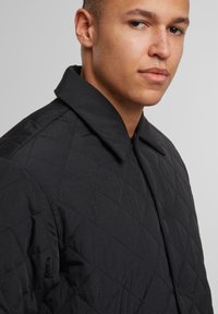 Giacca nera trapuntata caratterizzata da un ampio colletto camicia, tessuto testurizzato con motivi a diamante e un design snello.