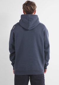 Sweat à capuche bleu foncé, fabriqué en tissu doux. Comprend une poche kangourou et un détail logo sur l'épaule gauche. Coupe décontractée.