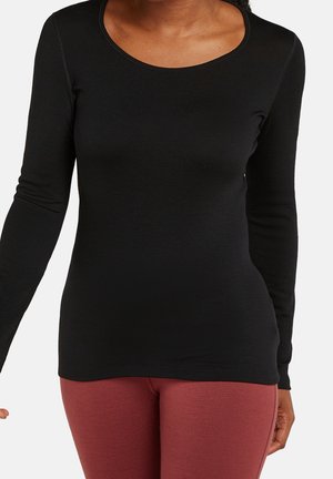 Femme portant un haut noir cintré à manches longues et encolure dégagée, avec un legging taille haute couleur rouille, debout devant un fond blanc.