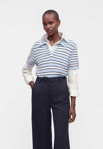 Femme aux cheveux courts portant un pull à manches courtes rayé bleu et blanc par-dessus une chemise blanche à col et un pantalon à fines rayures bleu marine.