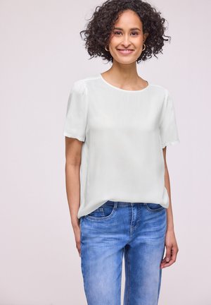 Smilende kvinde med krøllet hår iført guldringeøreringe, en hvid kortærmet bluse og blå jeans, stående mod en ensfarvet lys baggrund.