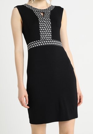 Czarna sukienka bez rękawów z geometrycznym wzorem przy dekolcie, talii oraz wycięciem na piersi, noszona przez stojącą modelkę.