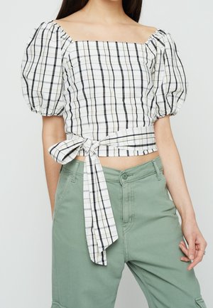 Mujer con un crop top de manga abullonada a cuadros en blanco y negro, con un lazo en la cintura, combinado con pantalones de talle alto en verde salvia.