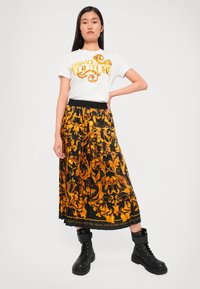 Bela majica s kratkimi rokavi z rumenim napisom "Versace Jeans Couture", v kombinaciji z črno krilom s cvetličnim vzorcem in naborki ter črnimi vojaškimi čevlji.