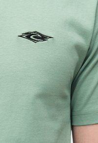 Ανοιχτό πράσινο κοντομάνικο πουκάμισο με μικρό μαύρο κεντημένο λογότυπο σε σχήμα διαμαντιού στο στήθος.