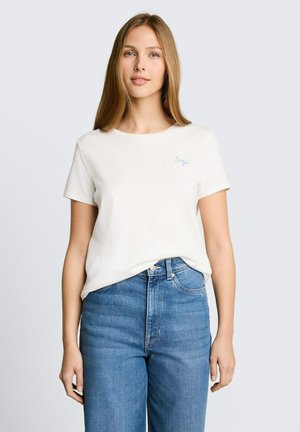 Junge Frau mit langen blonden Haaren, die ein weißes kurzärmeliges T-Shirt und hoch taillierte blaue Jeans trägt, vor einem einfarbigen Hintergrund stehend.