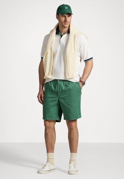 Polo shirt blanc avec des accents marine, short en coton vert, pull en mailles crème drapé sur les épaules, casquette verte et baskets blanches.