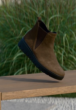 Bruine suède enkellaars met zwarte elastische zijpanelen en dikke zwarte rubberen zool die balanceert op een houten oppervlak buiten.