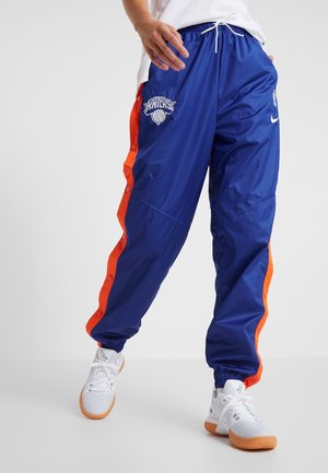 Personne portant un pantalon de basketball bleu des New York Knicks avec des bandes latérales orange et des chaussures de sport blanches à semelles en gomme.