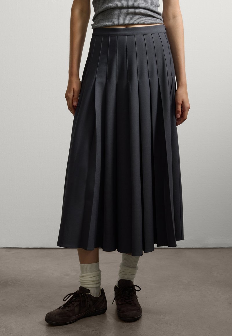 Donkergrijze geplooide rok met een brede tailleband, gemaakt van gladde stof. Draagt met bruine schoenen en crèmekleurige sokken; heeft een vloeiende vorm.