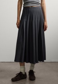 Gonna midi a pieghe grigio scuro con vita aderente, realizzata in tessuto morbido; indossata con scarpe scure e calzini chiari.
