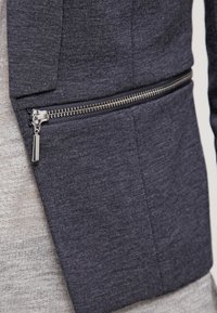 Mörkgrå texturerad jacka med en figursydd design, som har en synlig dragkedjeficka i silverfärgad hårdvara och kontrasterande ljusgrått tyg under.