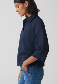 Haut bleu marine avec un col, des manches trois-quarts et une coupe décontractée. Le tissu semble lisse avec un design structuré.