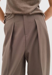 Pantaloni marroni a pieghe realizzati in tessuto liscio, con una vestibilità sartoriale e una trama morbida. Cuciture visibili lungo le cuciture.